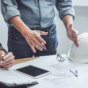 Engineering plans, a hard hat, and a tablet on a table, with a person's hands above them.