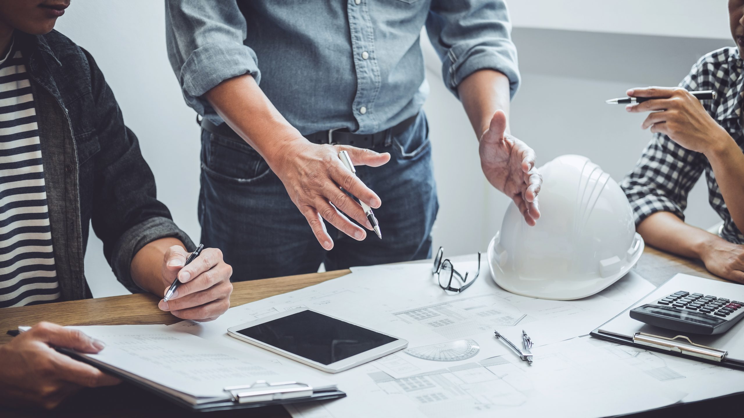 Engineering plans, a hard hat, and a tablet on a table, with a person's hands above them.